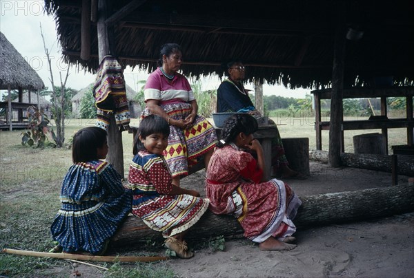 USA, Florida, Everglades, Independent Seminole Native American village. Family of women and young girls wearing traditional colourful patchwork dresses sitting under a Chickee hut.