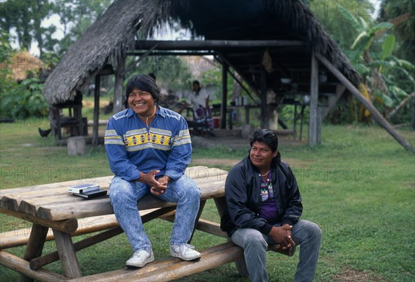 USA, Florida, Everglades, Independent Seminole Native American village. Two men named Danny and Billie sitting on a wooden bench smiling with a Chickee hut behind