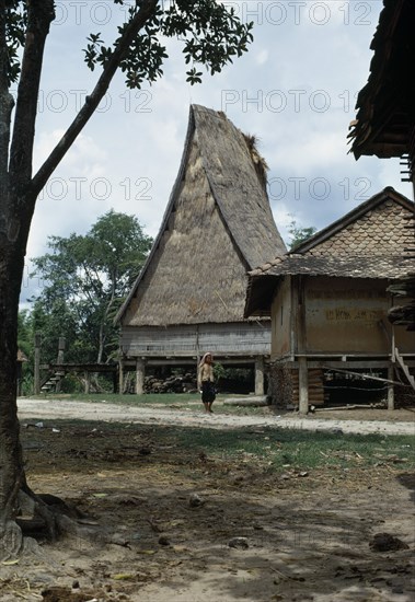 VIETNAM, Central Highlands, Kontum, Montagnard village. Woman walking along path in front of religious temple and family homes all thatched and built on stilts.
