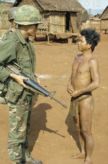 VIETNAM, Central Highlands, Plei Mrong, Vietnam War. Montagnard tribesman in Pleidok refugee camp with S.Vietnamese soldiers carrying guns standing over him. An-Khe Qui Nhon Pleiku BMT written on soldiers helmets.