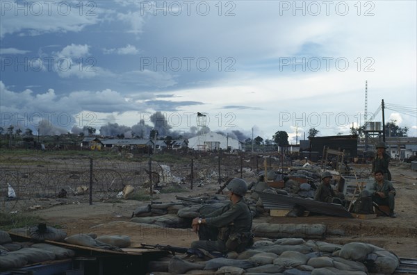 VIETNAM, Central Highlands, Kontum, Vietnam War. Siege of Kontum. American B-52 bombing behind Kontum. Montagnard soldiers sitting in camp within a barbed wire perimeter watching plumes of grey smoke rising into the sky