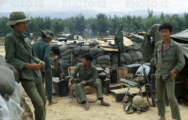 VIETNAM, Central Highlands, Kontum, "Vietnam War. Siege of Kontum. Montagnard soldiers gathered in base within a barbed wire perimeter. Young soldier sitting on a chair holding a medical syringe. Walls of sand bags, makeshift shelters, tools and artillery."