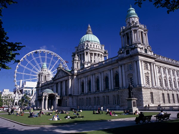 IRELAND, North, Belfast, People sitting or lying in the sun in the grounds of City Hall with The Wheel of Belfast part seen behind.