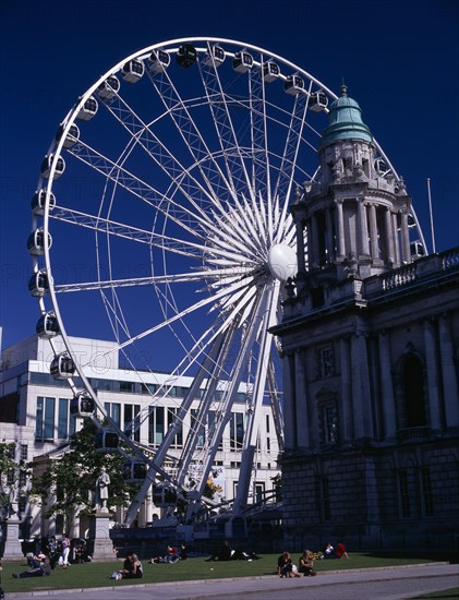 IRELAND, North, Belfast, People sitting or lying in the sun in the grounds of City Hall with The Wheel of Belfast part seen behind.