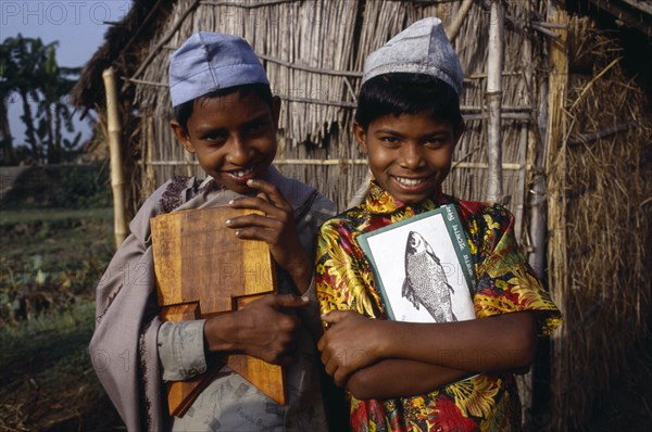 BANGLADESH, Shariatpur, "Three-quarter portrait of two, smiling young boys carrying books and writing board standing outside thatched hut."