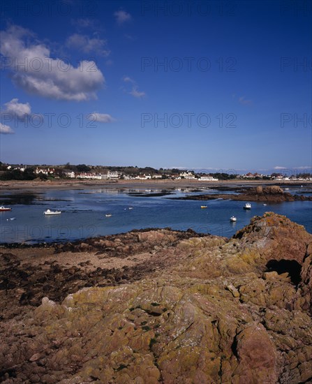 UNITED KINGDOM, Channel Islands, Jersey, Grouville. View across rocky foreshore with small boats on the sea towards Rocque Village