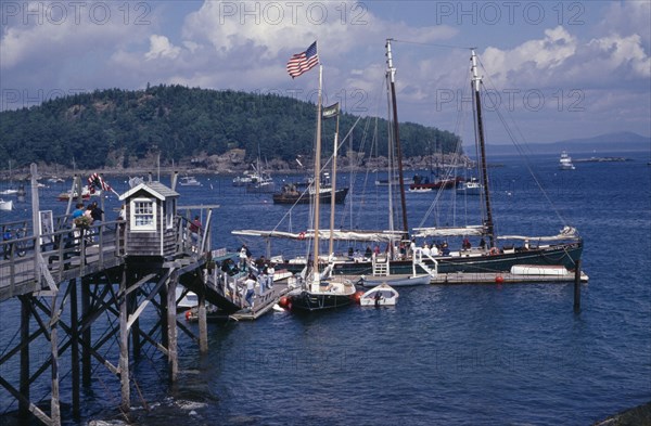 USA, Maine, Bar Harbour, "Wooden jetty with yacht moored at side, various boats on water and tree covered headland beyond."
