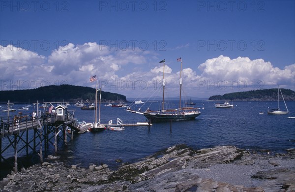 USA, Maine, Bar Harbour, "Wooden jetty with approaching yacht, various boats on water and tree covered headland beyond."