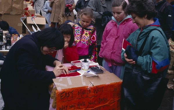 ENGLAND, London, Chinese New Year, Calligraphist writing Western names in Chinese during New Year celebrations in Chinatown.  Sign beside stall reads ‘East Meets West’.