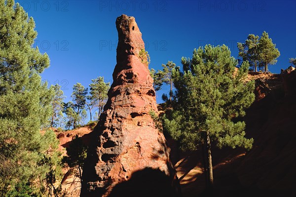 FRANCE, Provence Cote d’Azur, Le Sentier des Ocres (The Ochre Footpath) , "A tall thin, eroded rock pinnacle in the area known as the Needle Cirque part circled by trees against cloudless blue sky."