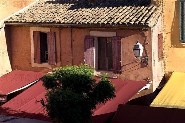 FRANCE, Provence Cote d’Azur, Vaucluse, "Roussillon.  Provencal colours in the town centre, exterior facade of building with tiled roof, red awning and pale ochre and orange walls."