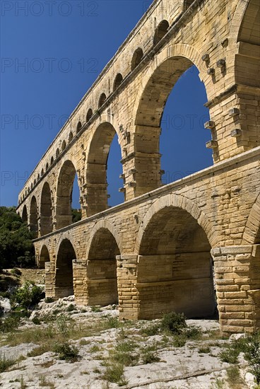 FRANCE, Provence Cote d’Azur, Gard, Pont du Gard.  Angled view of three tiers of arches of Roman aqueduct from the west side in glowing evening light.