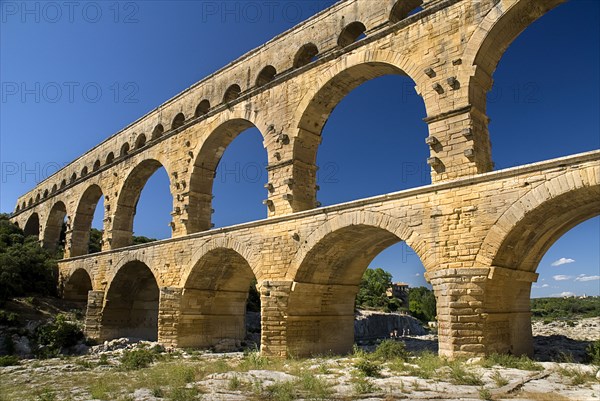 FRANCE, Provence Cote d’Azur, Gard, Pont du Gard.  Angled view of Roman aqueduct from the west side in glowing evening light showing three tiers of continuous arches against cloudless blue sky.