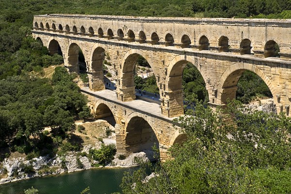 FRANCE, Provence Cote d’Azur, Gard, Pont du Gard Roman aqueduct from a high vantage point on the western side showing three tiers of continuous arches spanning river and visitors on bridge.