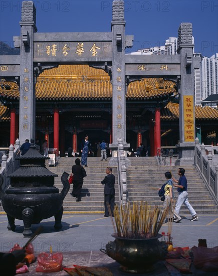 CHINA, Hong Kong, Kowloon, "Wong Tai Sin taoist temple established in 1921.  Visitors on flight of steps to entrance built in traditional Chinese style with red pillars and pagoda style roof.   Incense, offerings and burner in foreground. "