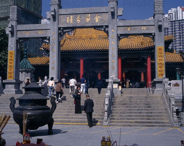 CHINA, Hong Kong, Kowloon, "Wong Tai Sin taoist temple established in 1921.  Visitors on flight of steps to entrance built in traditional Chinese style with red interior pillars and pagoda style roof.   Incense, offerings and burner in foreground. "