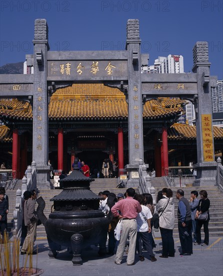 CHINA, Hong Kong, Kowloon, Wong Tai Sin taoist temple established in 1921.  Visitors gathered beside incense burner at foot of flight of steps to entrance built in traditional Chinese style with red interior pillars and pagoda style roof.