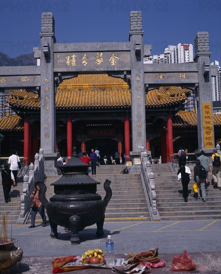 CHINA, Hong Kong, Kowloon, "Wong Tai Sin taoist temple established in 1921.  Visitors on flight of steps to entrance built in traditional Chinese style with red interior pillars and pagoda style roof.   Incense, burner and offerings in foreground. "