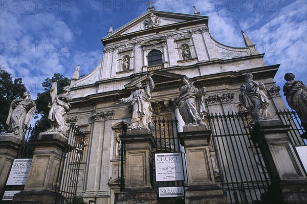 POLAND, Krakow, Exterior facade of Church of Saints Peter and Paul with  statues of the twelve disciples and religious iconography and sign detailing times of Mass services on ironwork gate.