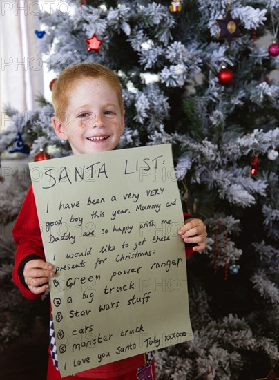 RELIGION, Festivals, Christmas, Young smiling red headed boy standing in front of a decorated Christmas Tree holding up the Santa List he has written on Christmas Eve