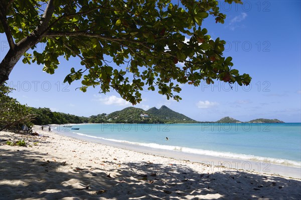 WEST INDIES, Grenada, Carriacou, The calm clear blue water breaking on Paradise Beach in L'Esterre Bay with Cistern Point and the Sisiter Rocks in the distance. A small number of people on the beach and in the water