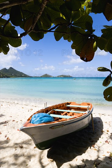 WEST INDIES, Grenada, Carriacou, The calm clear blue water breaking on Paradise Beach in L'Esterre Bay with Cistern Point and the Sister Rocks in the distance. A small fishing boat sits on the sandy shoreline out of the water