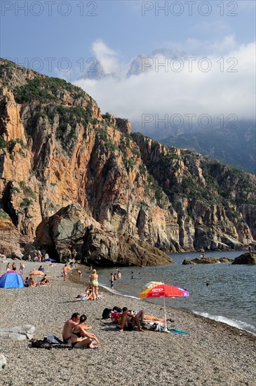 FRANCE, Corsica, Golfe Di Porto, "Plage De Bussaglia, sandy beach with sunbathers & umbrellas. Rocky cliffs & cloud on hills"