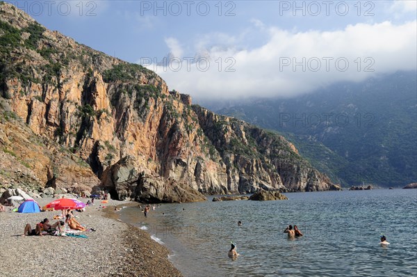 FRANCE, Corsica, Golfe Di Porto, "Plage De Bussaglia, sandy beach with sunbathers & umbrellas. Rocky cliffs & cloud on hills"