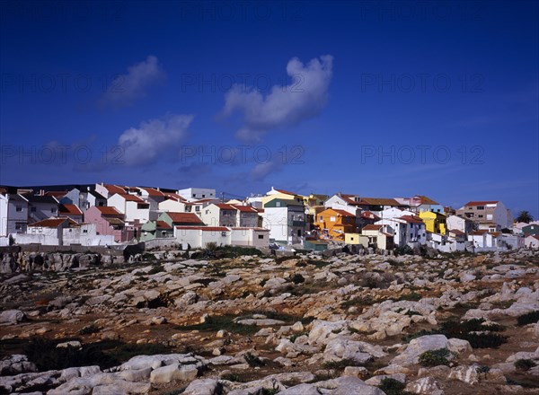 PORTUGAL, Estremadura, Ribatejo, Peniche. Atlantic Ocean town. View across low cliffs and rocky foreshore towards colourful painted houses with red tiled roofs on the south side of the town