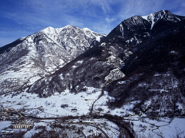 SPAIN, Catalonia, Lleida, Pyrenees. Elevated view north east from the route to Col du Portillon looking over the rooftops of  the town of Ville Bossot. Snow covered mountains including Montlude 2518 metres 8246 feet and Montanna d’Uishera 2342 metres 7670 feet. Valle Garone river seen on the valley below.