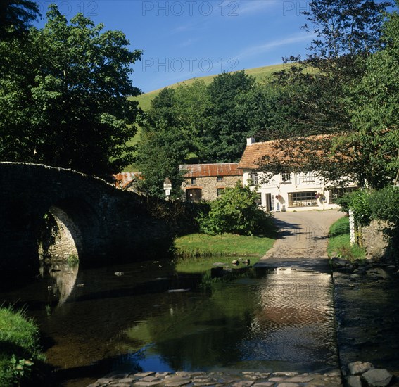 ENGLAND, Devon, Exmoor, Malmsmead on Badgworthy Water. Arched stone bridge and ford across river leading to ‘Lorna Doone Farm’ shop White painted building with tiled roof  and outbuildings.