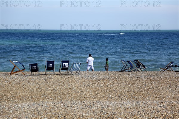 ENGLAND, East Sussex, Eastbourne, Man and young boy on the beach with deck chairs blowing in the wind. Speed passing by in the distance.