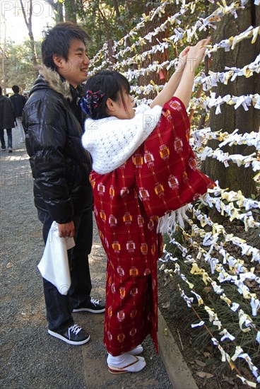 JAPAN, Honshu, Tokyo, "Jingumae - at Meijijingu shrine, couple boy and girl early twenty years old, girl in traditional kimono and wrap, boy in down jacket, jeans, sneakers, tie omikuji New years prediction to a fencr."