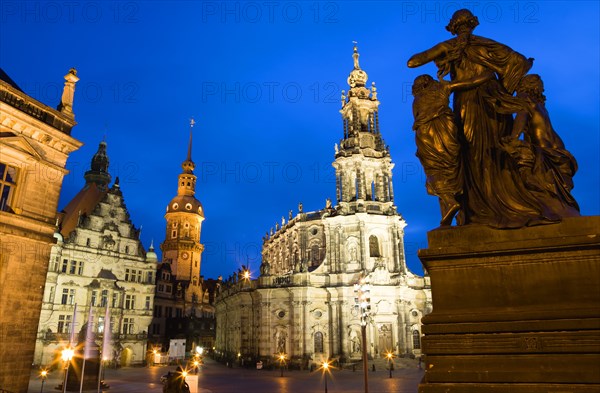 GERMANY, Saxony, Dresden, The 18th Century Hofkirche Catholic Cathedral of Saint Trinitatis and the illuminated tower of the Residenzschloss and the Grunes Gewelbe or Green Vault a museum that is part of Dresden Castle with a statue of a woman and children in the foreground at sunset.