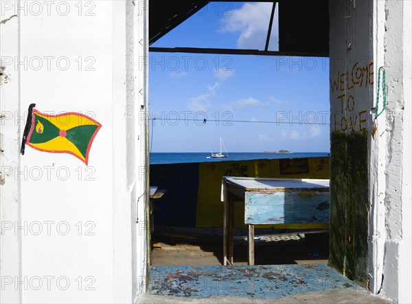 WEST INDIES, Grenada, Carriacou, Hillsborough Derelict building in the main street with the Grenadian flag and Welcome To Love painted on the walls with a yacht at anchor in the harbour beyond the ruined building.