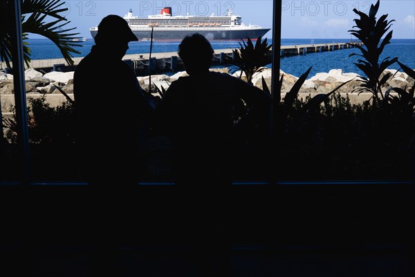 WEST INDIES, Grenada, St Georges, Tourists in the Cruise Ship Terminal looking out through the glass window towards the Queen Mary 2 liner moored offshore.