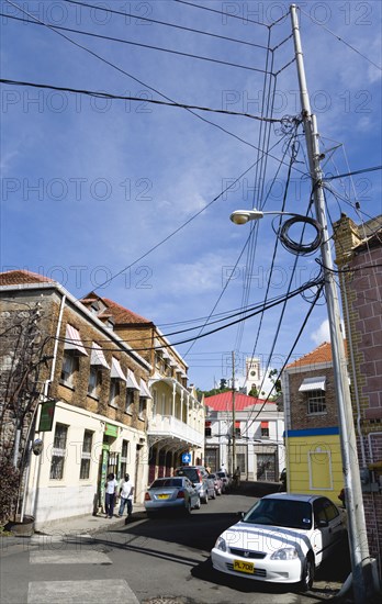 WEST INDIES, Grenada, St Georges, People walking down a side street lined with parked cars in the capital with overhead electricity power and telephone cables.