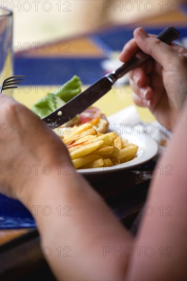 WEST INDIES, Grenada, St George, Sunburnt white caucasian woman eating a plate of potato chips and tomato ketchup with a knofe and fork.