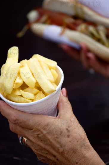 ENGLAND, West Sussex, Findon, Findon village Sheep Fair Elderly lady holding a polystyrene mug full of potato fries in one hand and a hot dog with tomato ketchup and onions in the other.