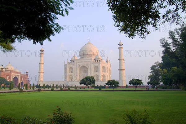 INDIA, Uttar Pradesh, Agra, Taj Mahal at sunrise one of the wonders of the world.  Exterior facade with domed roof and minarets part framed by trees.