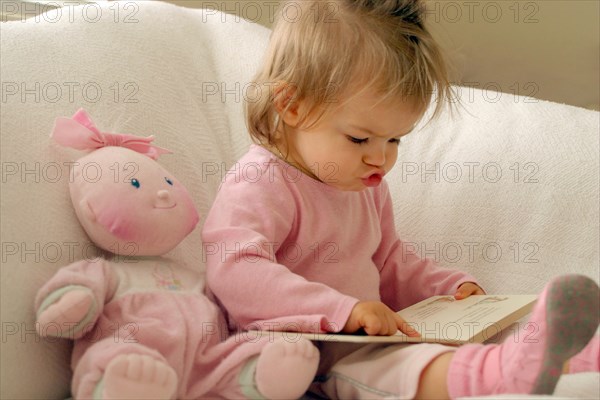 EDUCATION, Children, Reading, Infant girl dressed in pink reading book whilst mouthing the words and following text on the page with her finger.  Pink doll propped up on chair beside her.