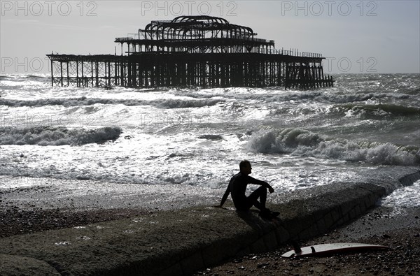 ENGLAND, East Sussex, Brighton, "Seafront, man sat with surf board on goyne in front of the burnt out shell of the former West Pier. "