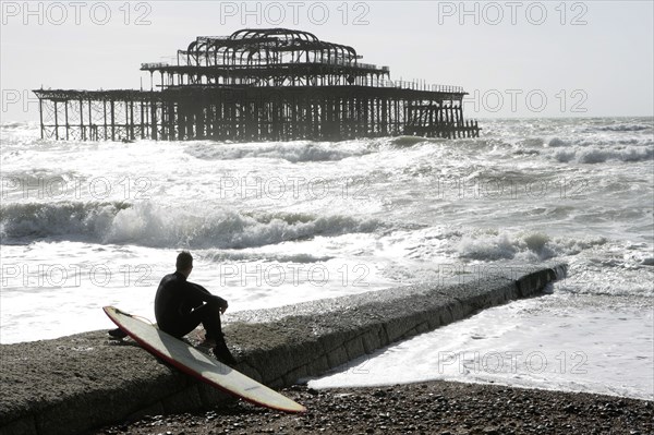 ENGLAND, East Sussex, Brighton, "Seafront, man sat with surf board on goyne in front of the burnt out shell of the former West Pier. "