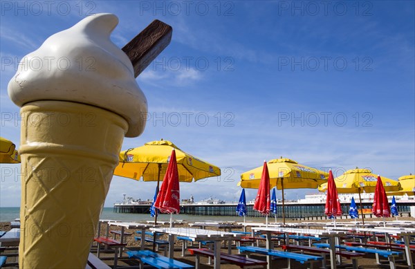 ENGLAND, East Sussex, Brighton, The Pier at low tide with shingle pebble beach and sunshade umbrellas by empty tables on the seafront with a fibreglass ice cream cone in the foreground.