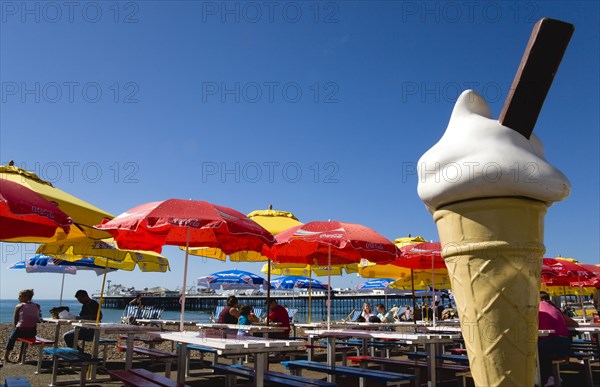 ENGLAND, East Sussex, Brighton, The Pier with people under sunshade umbrellas by tables on the seafront with a fibreglass ice cream cone in the foreground.