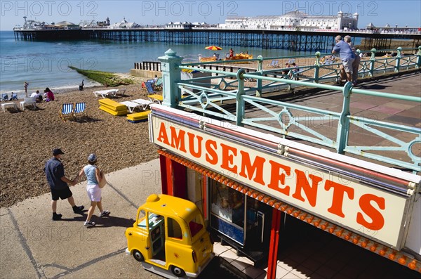 ENGLAND, East Sussex, Brighton, The Pier with people on the shingle pebble beach walking along the promenade and looking out to sea from above an amusements arcade on the seafront.