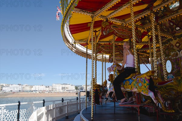 ENGLAND, East Sussex, Brighton, The Pier with a happy laughing young girl on a traditional Victorian galloping horses fairground carousel roundabout ride with the beach and seafront buildings in the distance.