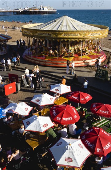 ENGLAND, East Sussex, Brighton, People sitting under sun shade umbrellas at tables on the promenade outside bars and restaurants with a traditional Victorian galloping horses carousel fairground roundabout and Brighton Pier with people on the shingle pebble beach beyond.