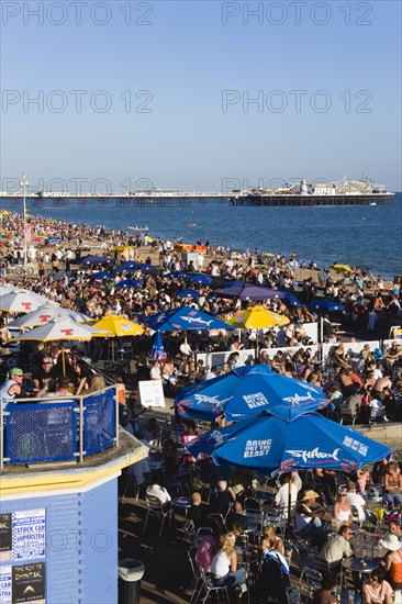ENGLAND, East Sussex, Brighton, People sitting under sun shade umbrellas at tables on the promenade outside bars and restaurants with Brighton Pier and people on the shingle pebble beach beyond.
