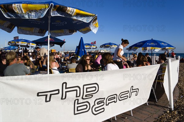 ENGLAND, East Sussex, Brighton, People sitting under sun shade umbrellas at tables on the promenade outside The Beach Bar with The Brighton Pier beyond.